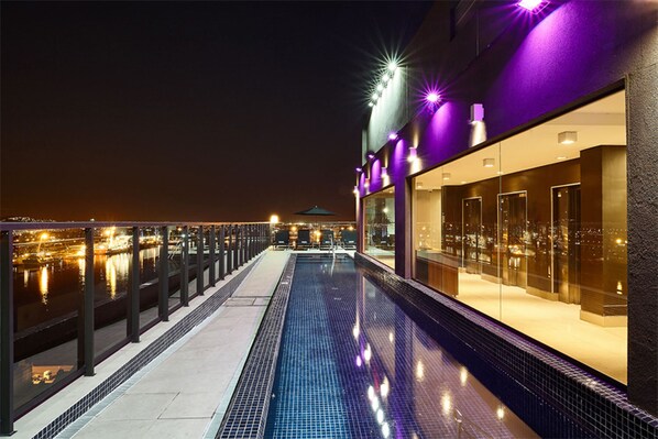 Vue nocturne d’une piscine sur le toit, avec des garde-corps en verre, donnant sur la ville, et d’un bâtiment illuminé de lumières violettes.