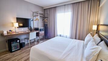 Habitación de hotel con cama, escritorio, televisión, refrigerador pequeño y ventana con cortinas.