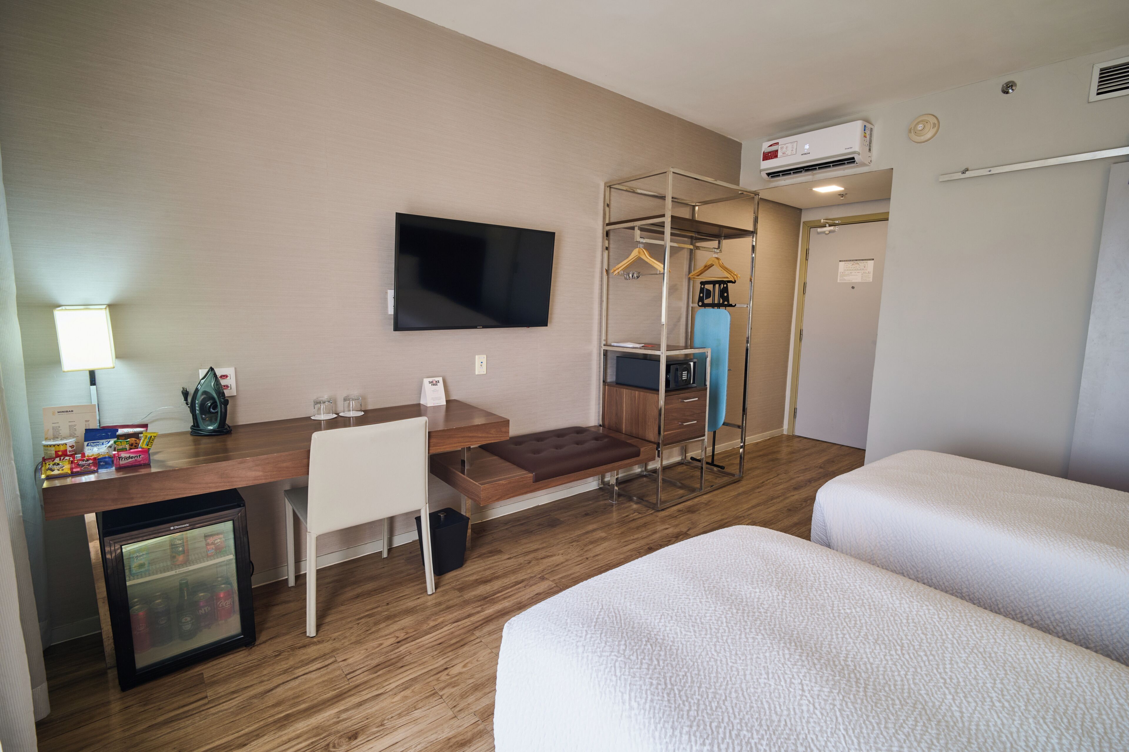 Habitación de hotel con una cama, un escritorio con computadora, televisor, mini refrigerador, armario y perchero.