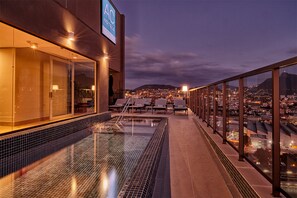 Un espace piscine sur le toit, doté de rampes en verre, de chaises longues et offrant une vue sur la ville au crépuscule.