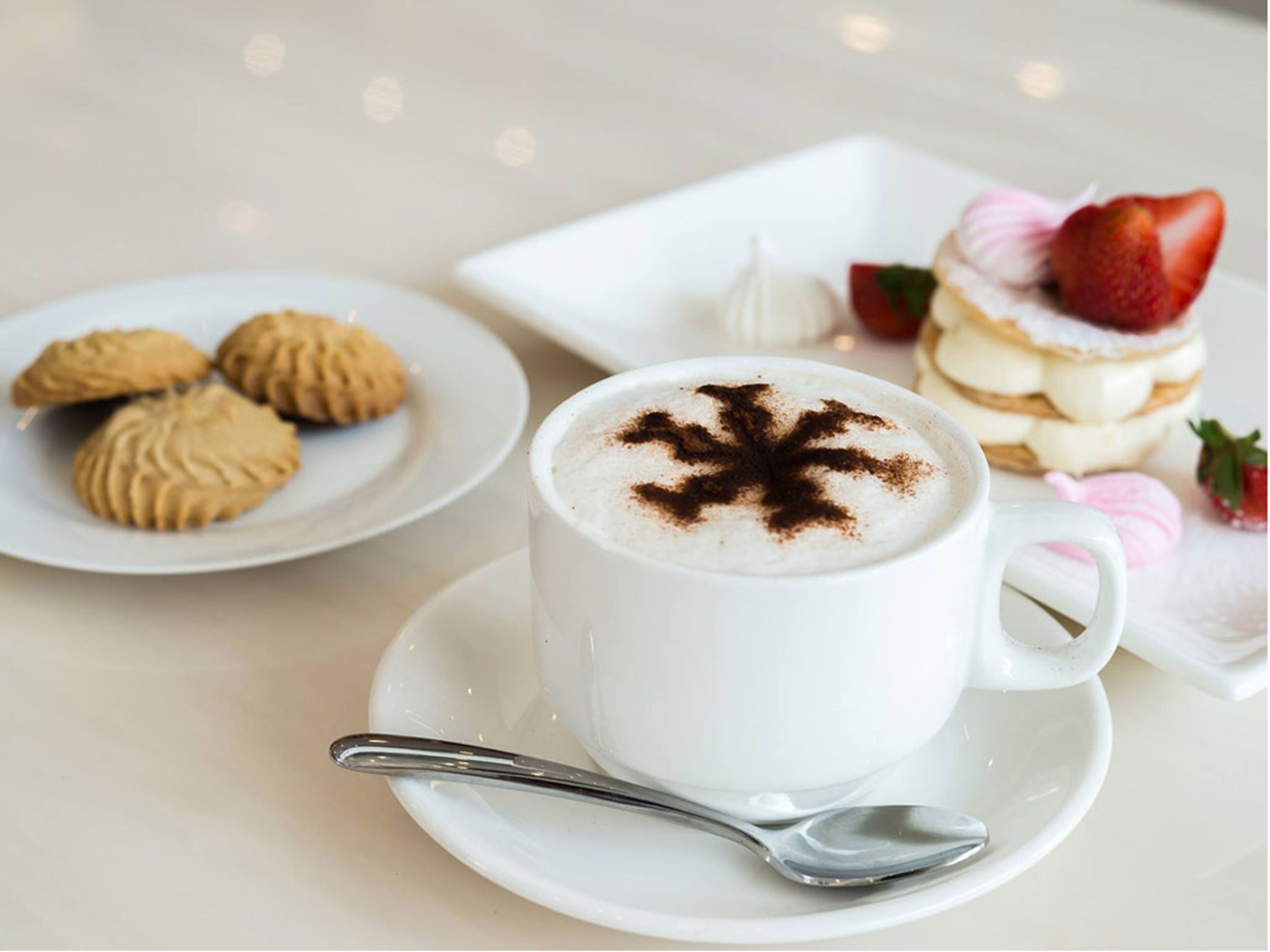 A cup of coffee with a chocolate design on top, a plate of cookies, and a dessert with strawberries.