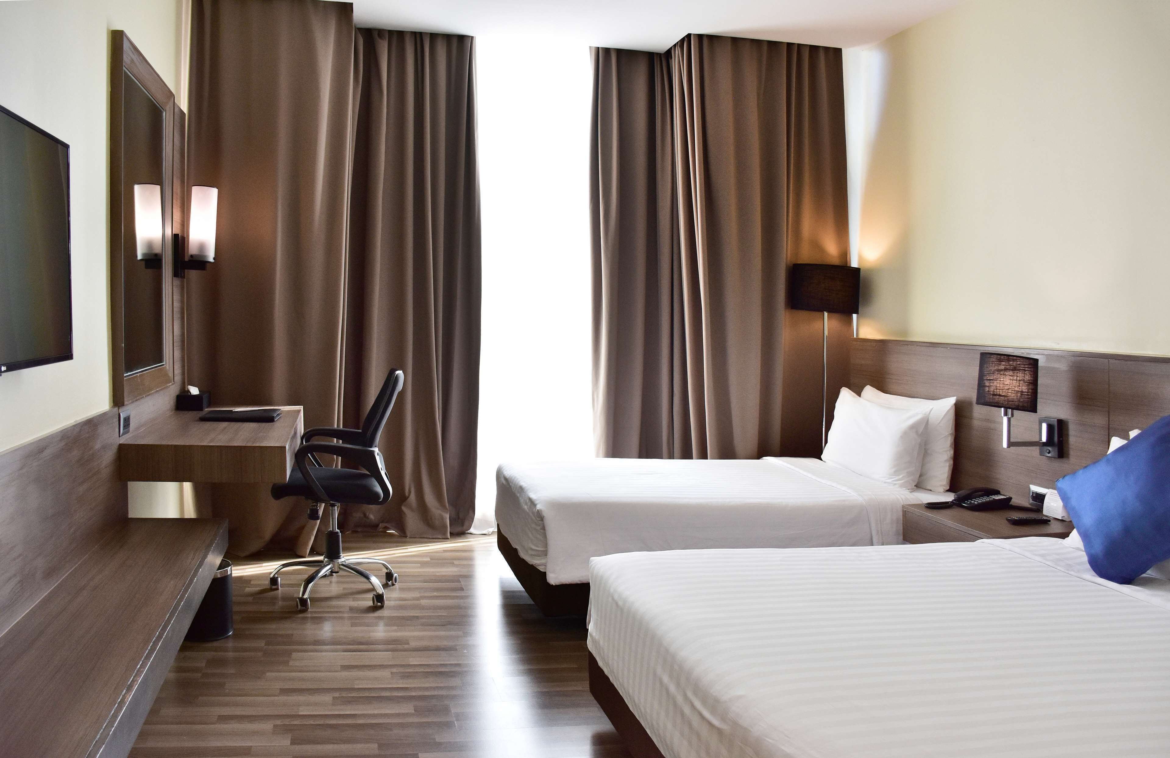 A hotel room with two beds, a desk, a chair, a TV, and a nightstand with a lamp.