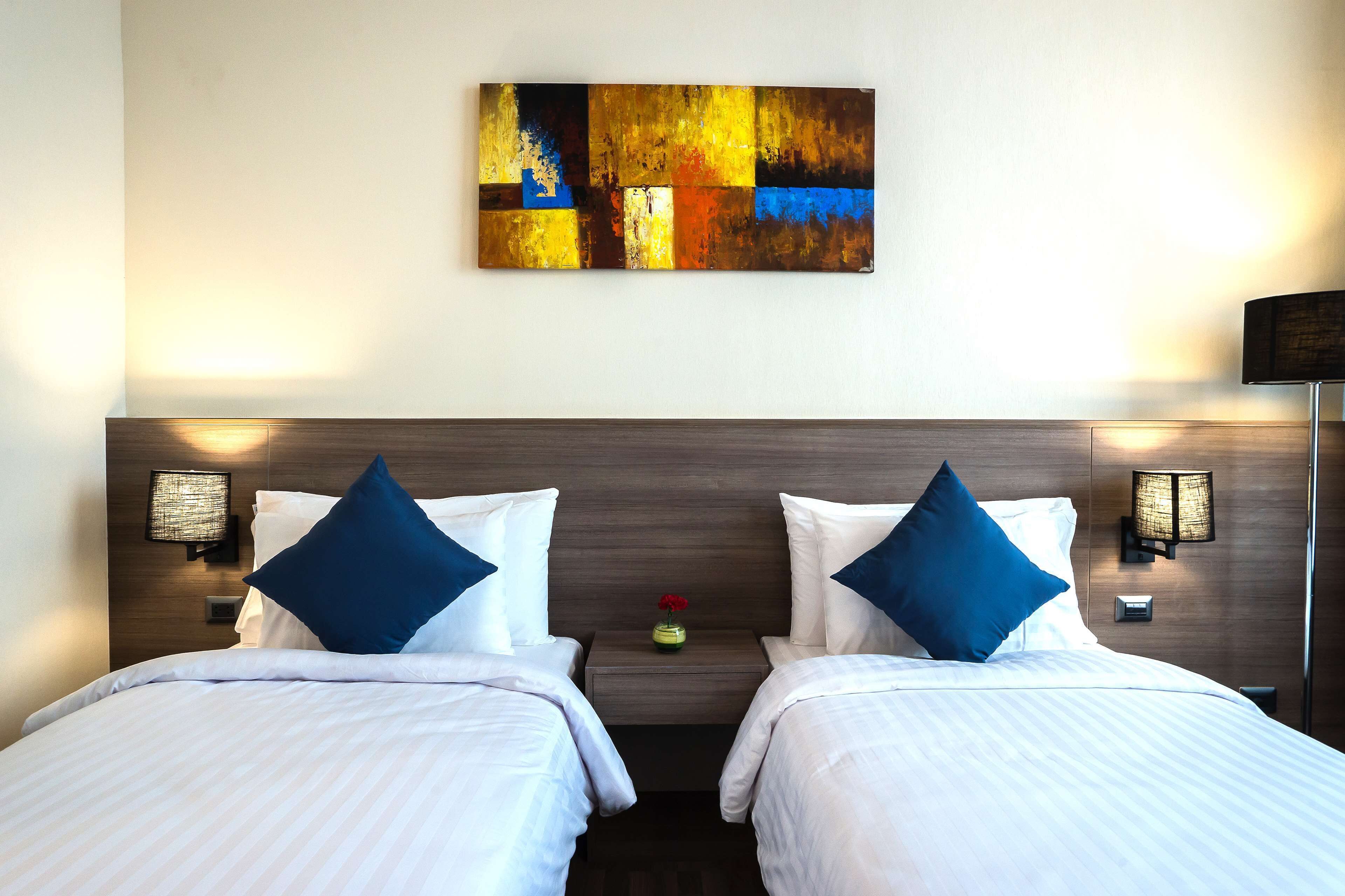 A hotel room with two beds, a wooden headboard, a wall-mounted abstract painting, bedside lamps, and a small table with a vase.
