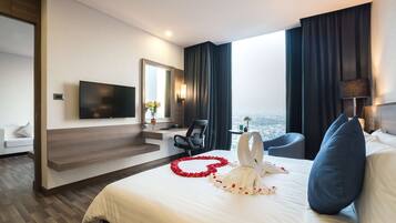 A hotel room with a large bed, a flat-screen TV, a desk with a chair, a sofa, and a view of the city.