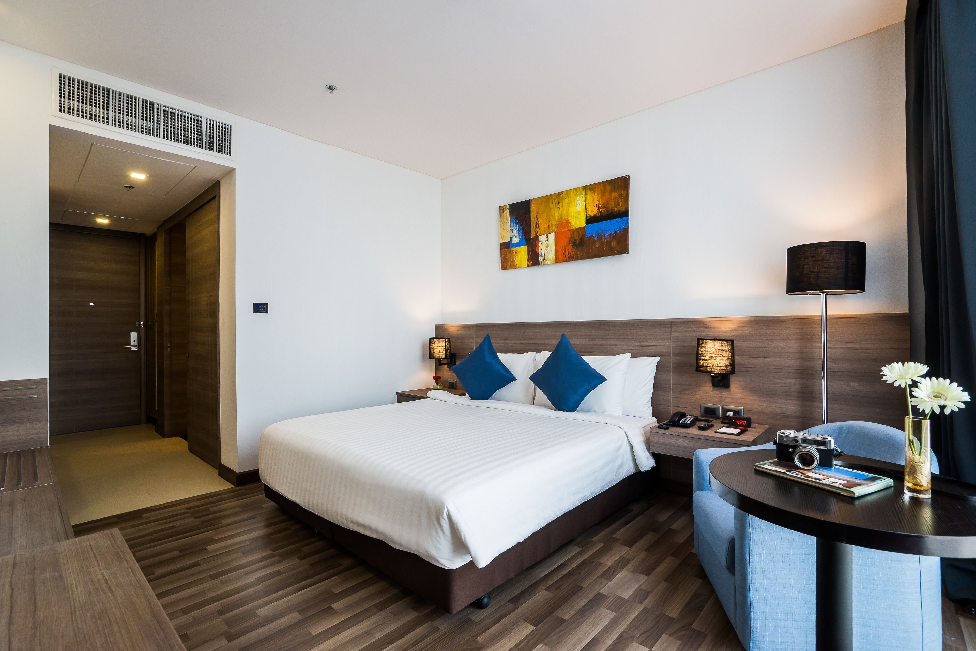 A modern hotel room with a large bed, a desk with a lamp, and a painting on the wall.