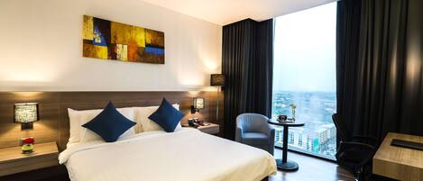 A hotel room with a large bed, a desk, a chair, a small table with a vase, and a city view.