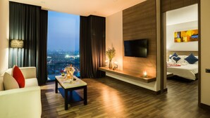Quarto de hotel moderno com vista para a cidade, televisão de tela plana, área de refeições e cama aconchegante com almofadas decorativas.