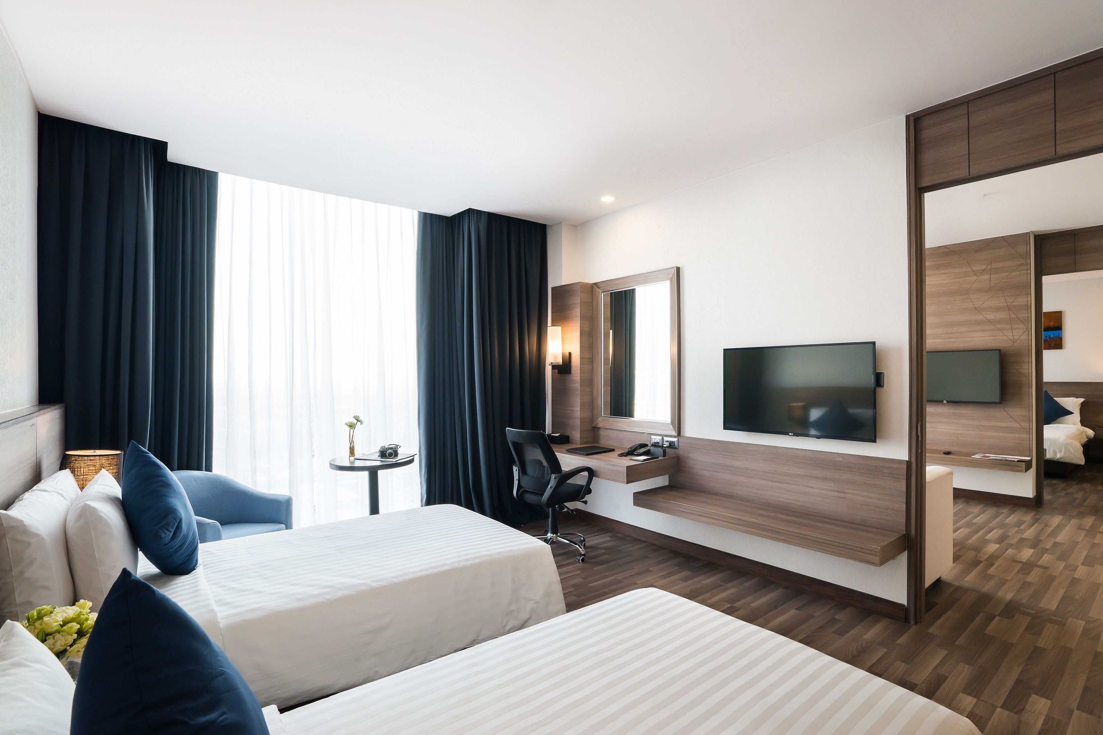 A modern hotel room with two beds, a desk, a chair, a TV, and a bathroom visible through a sliding door.