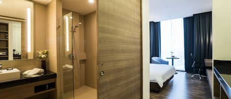 Una habitación de hotel moderna con ducha de vidrio, pared de paneles de madera, una cama grande, un escritorio y un baño con tocador y espejo.