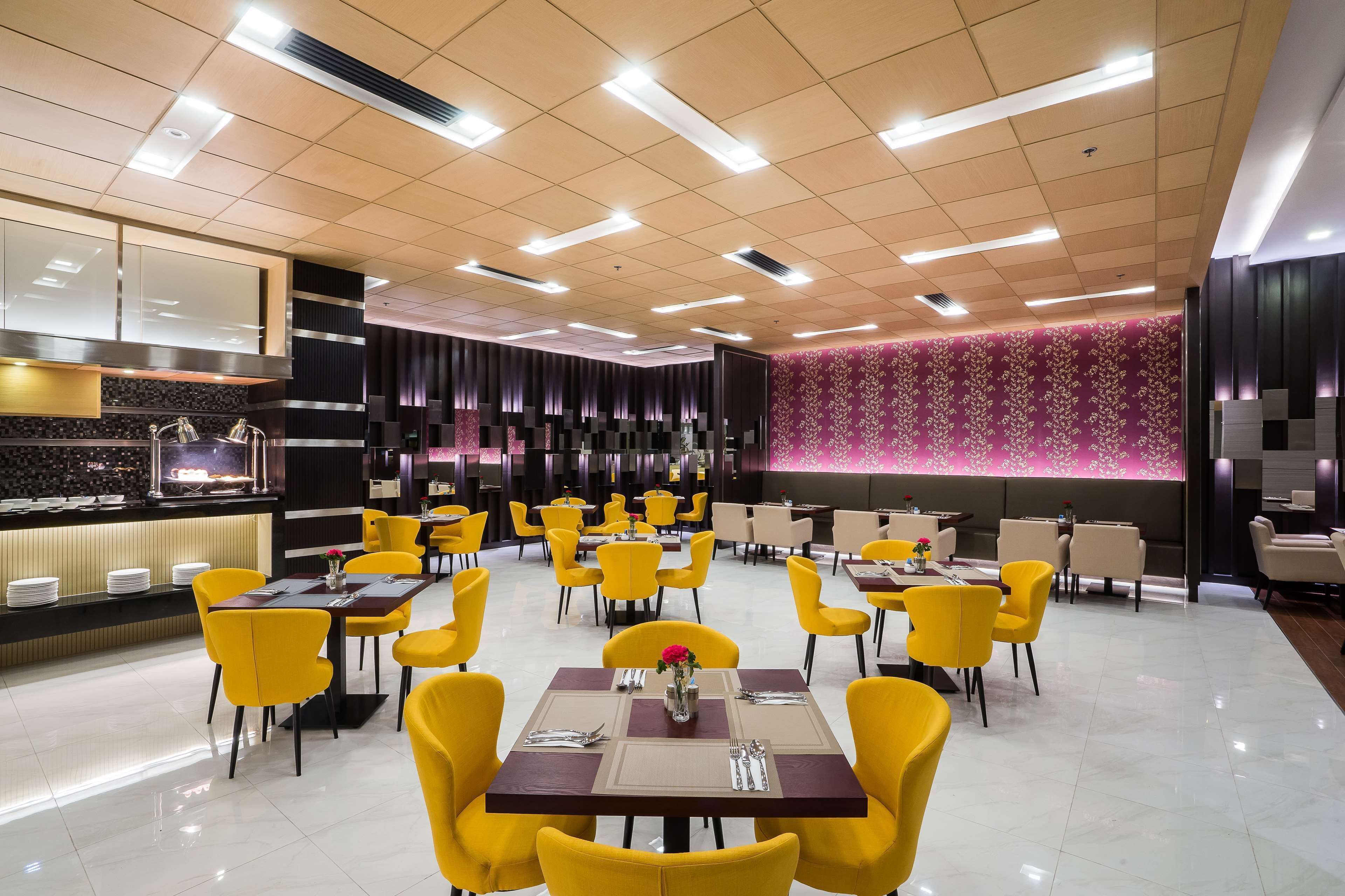 Ein modernes Restaurant mit gelben Stühlen und Tischen, einer Theke mit Tellern und einer Wand mit einem rosa Muster.