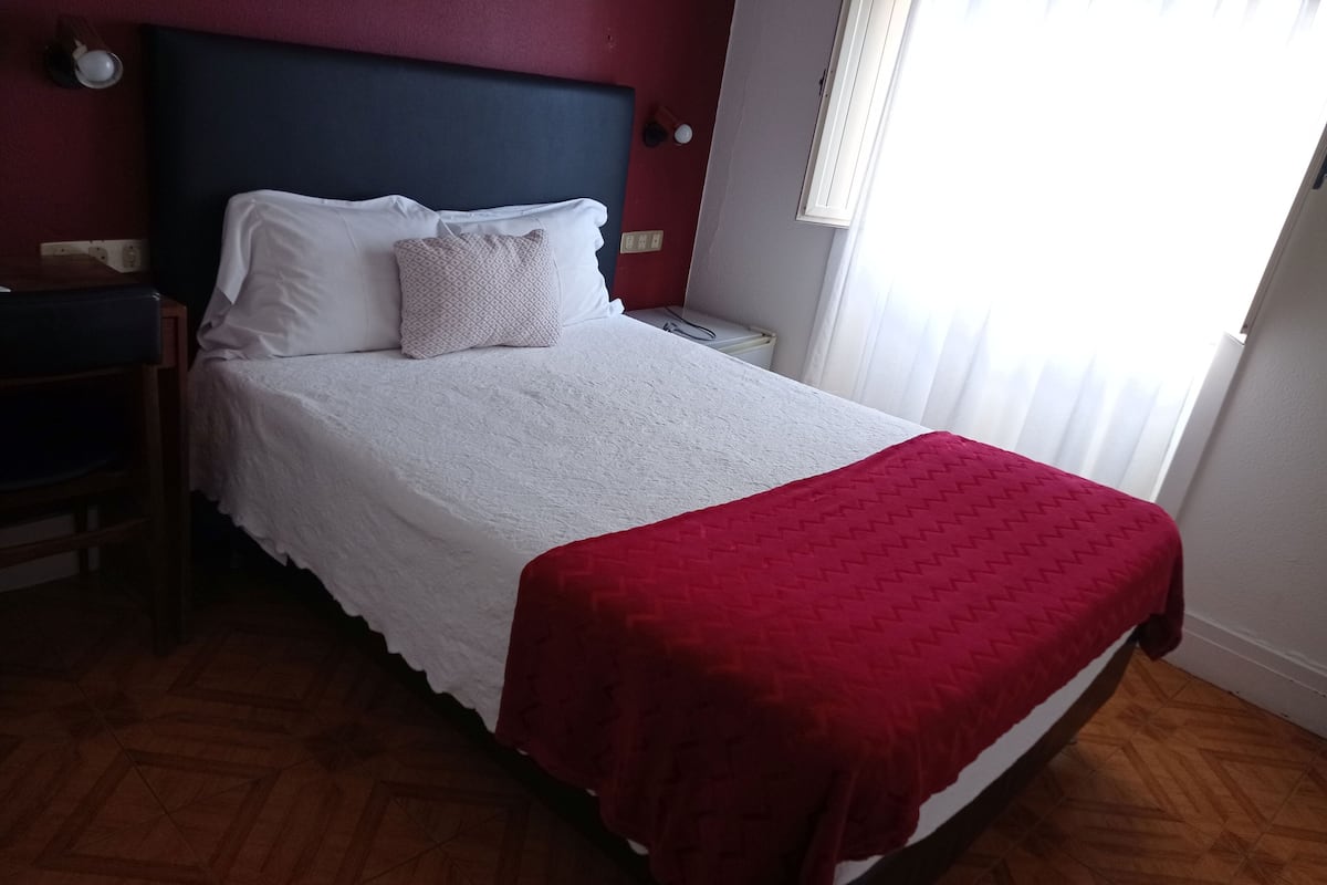 A neatly made bed with a white duvet and a red blanket, a wooden nightstand with a lamp, and a window with blinds.