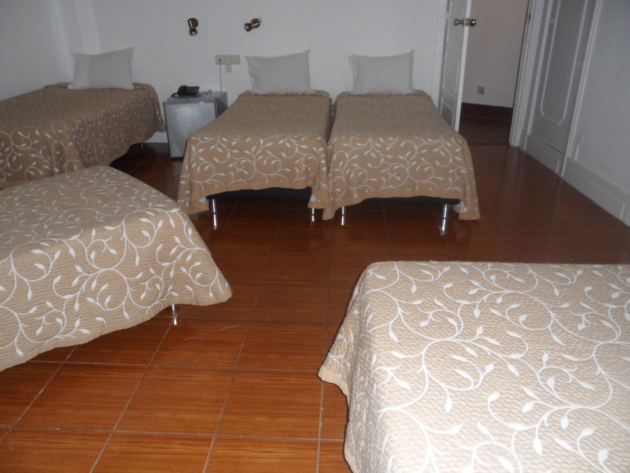 Une chambre d’hôtel avec trois lits, chacun recouvert d’une couvre-lit à motifs, une table de chevet avec une lampe et une prise électrique au mur.