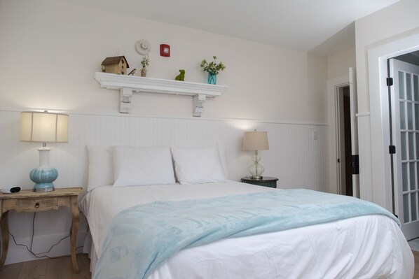 A bedroom with a bed, bedside tables, a wall-mounted shelf with decorative items, and a door to the right. - Marginal Way House Hotel (Ogunquit)