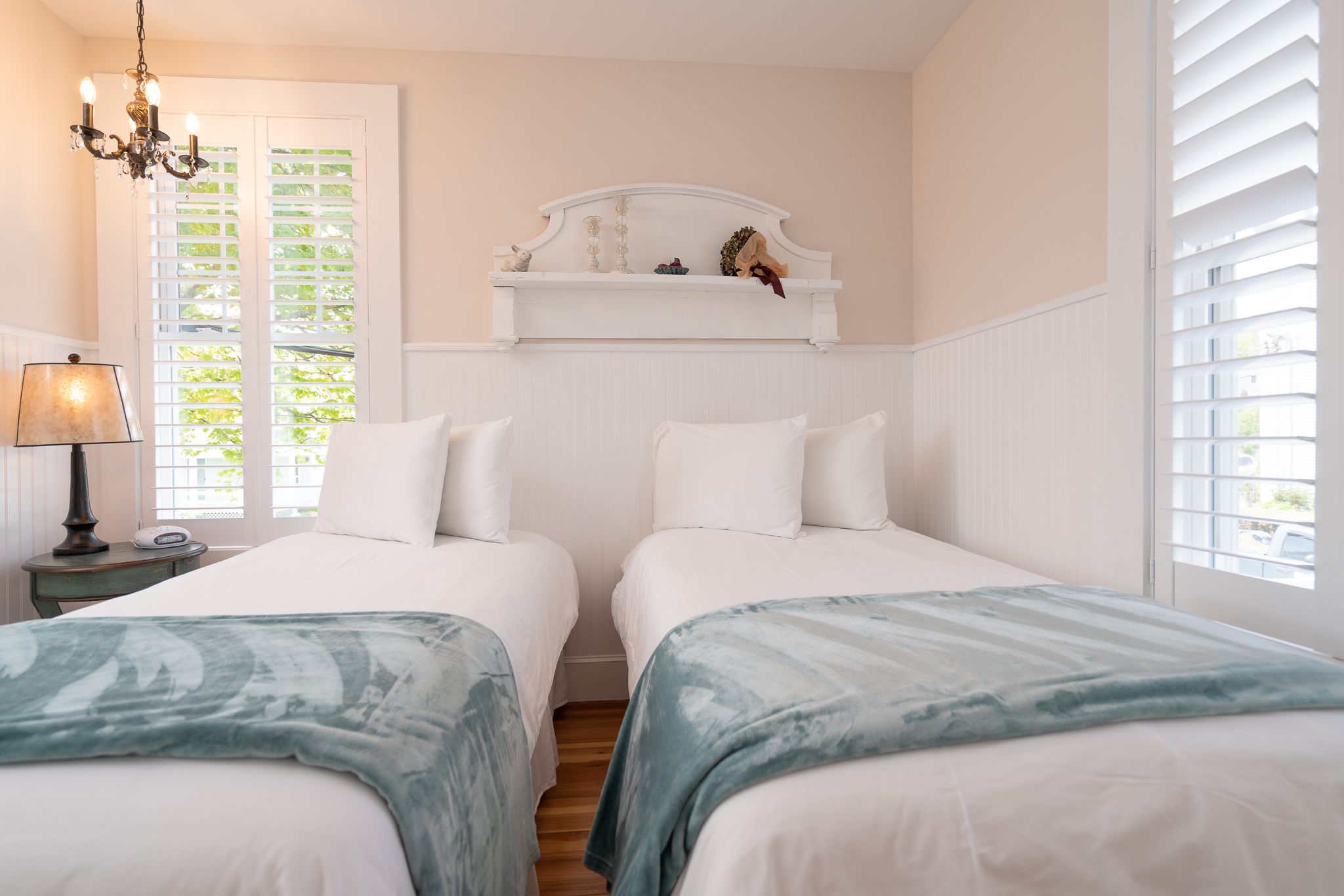 Quarto com duas camas, mesa de cabeceira com abajur, cabeceira decorativa e vista para o verde pela janela.