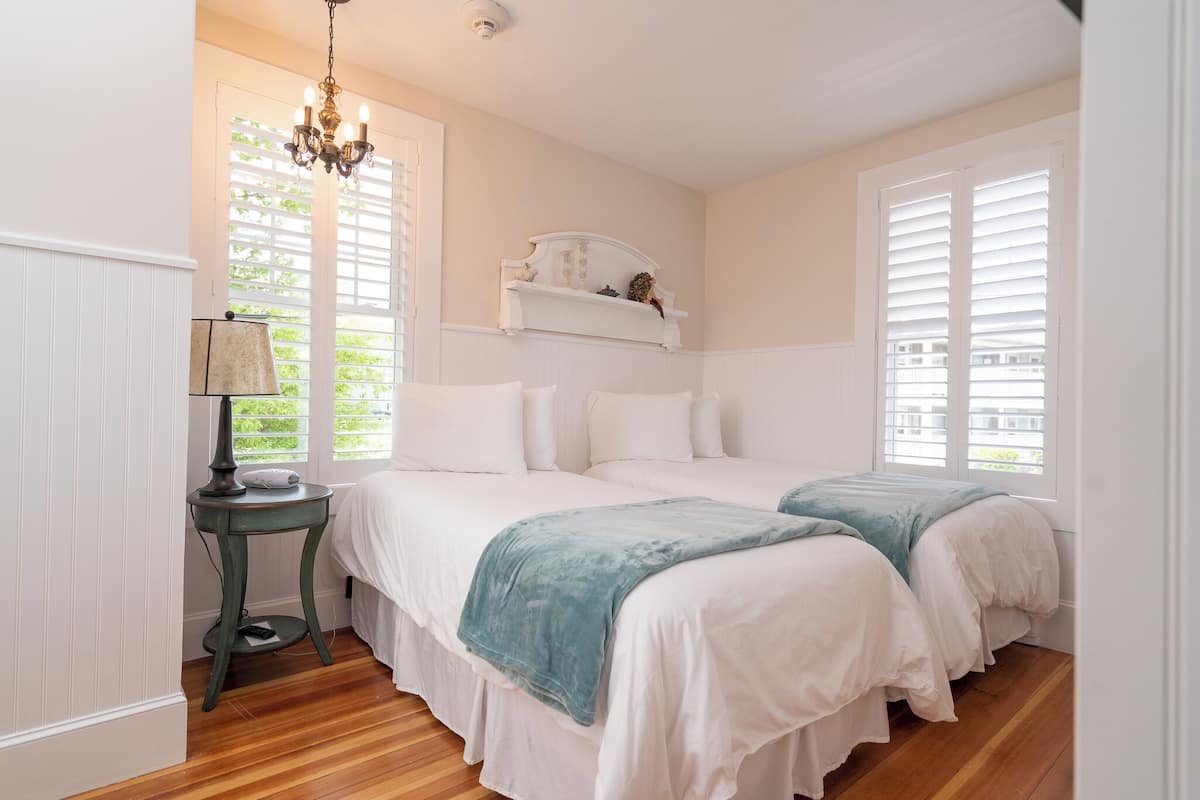 A bedroom with two beds, a bedside table with a lamp, a white headboard, and a window with blinds.