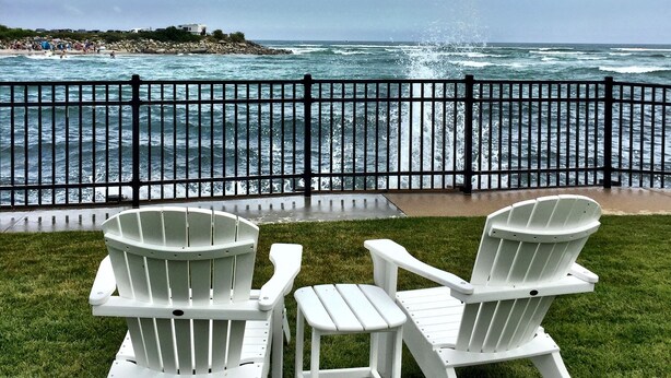 Two white Adirondack chairs facing a rocky coastline with a view of the ocean.