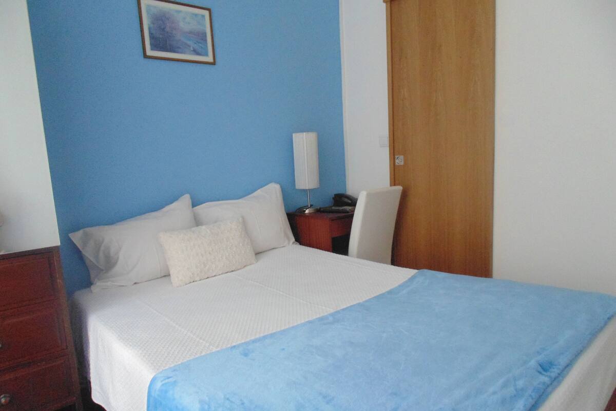A neatly made bed with white bedding and a blue accent, a wooden nightstand with a lamp, a chair, and a wooden door.