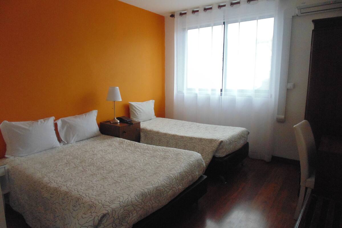 A hotel room with two beds, a nightstand with a lamp, and a window with curtains.