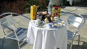 A round table with a white tablecloth, surrounded by metal chairs, set outdoors with a concrete floor and some greenery in the background. - Hotel Porto Rico (Porto)