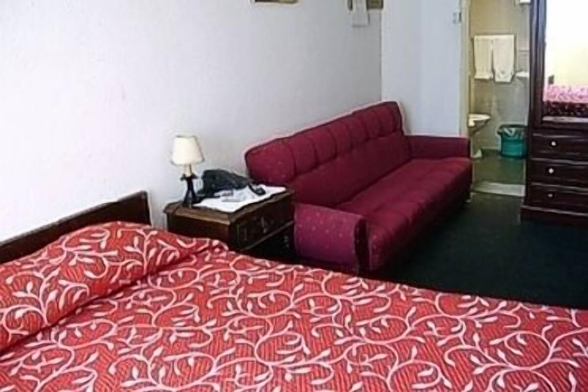 A hotel room with a bed, a red sofa, a nightstand with a lamp, and a dresser.