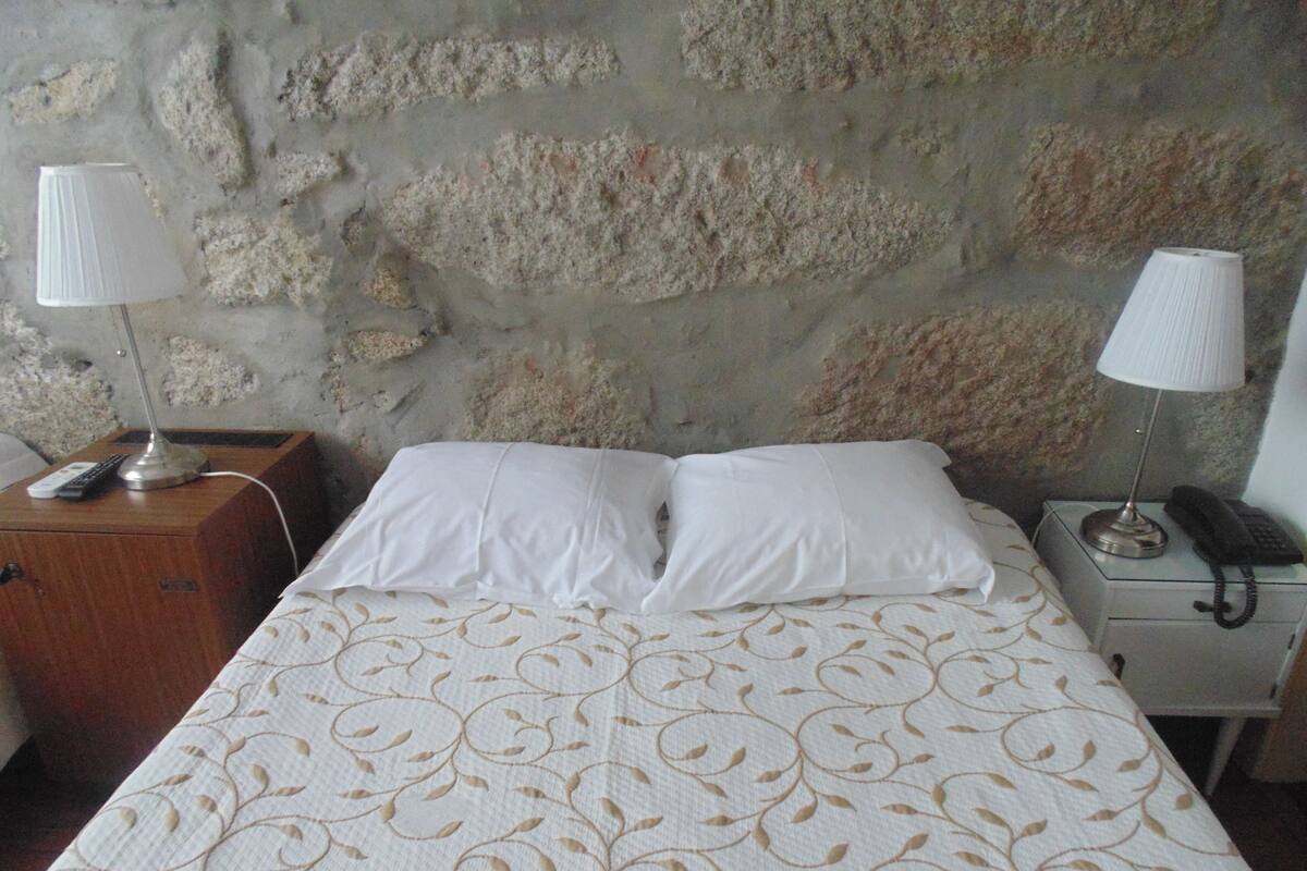 A neatly made bed with a patterned bedspread, two bedside lamps, a bedside table with a lamp and a phone, and a wall with a rough stone texture.