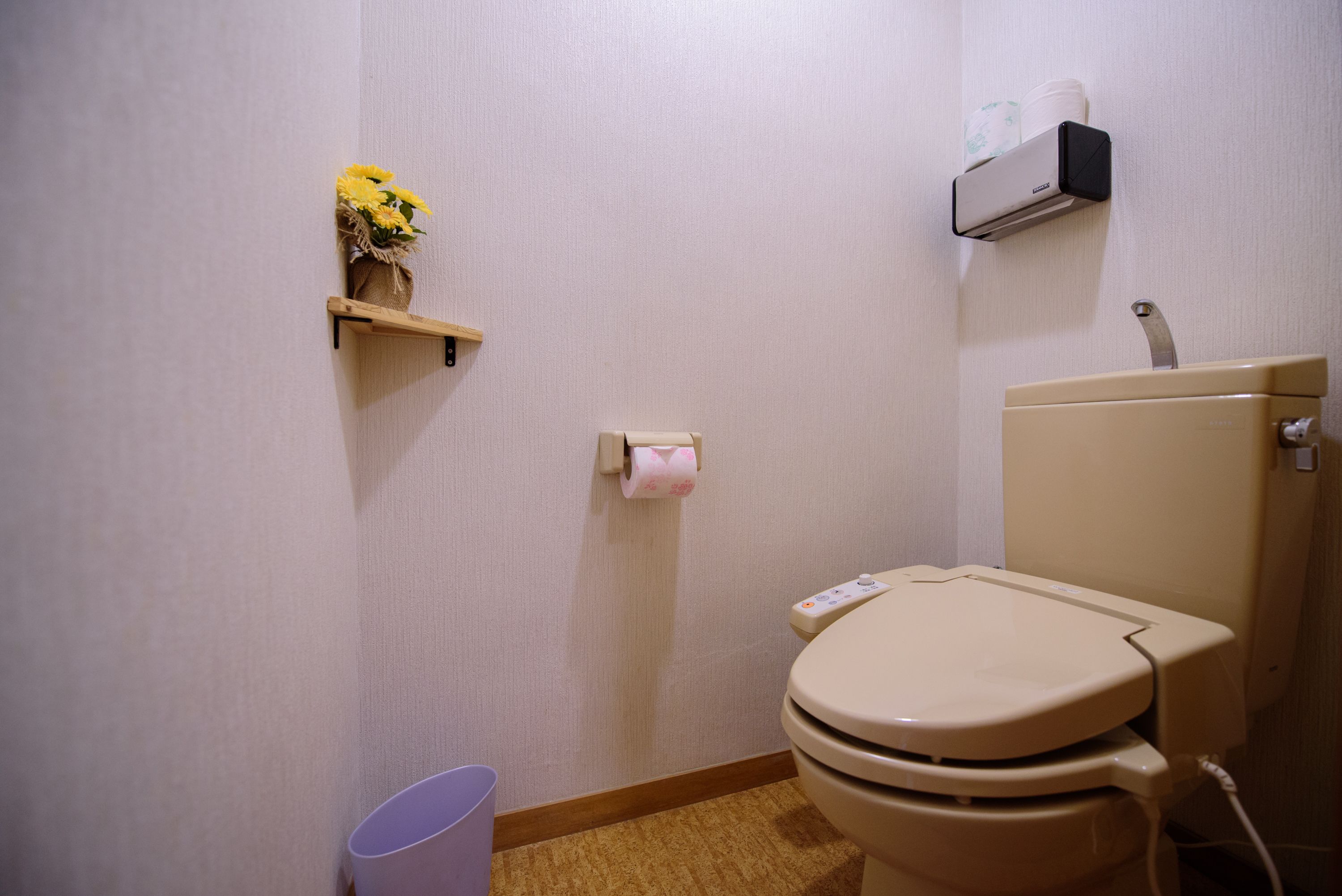 Un baño pequeño con inodoro, un dispensador de papel higiénico, un bote de basura y un estante con flores.