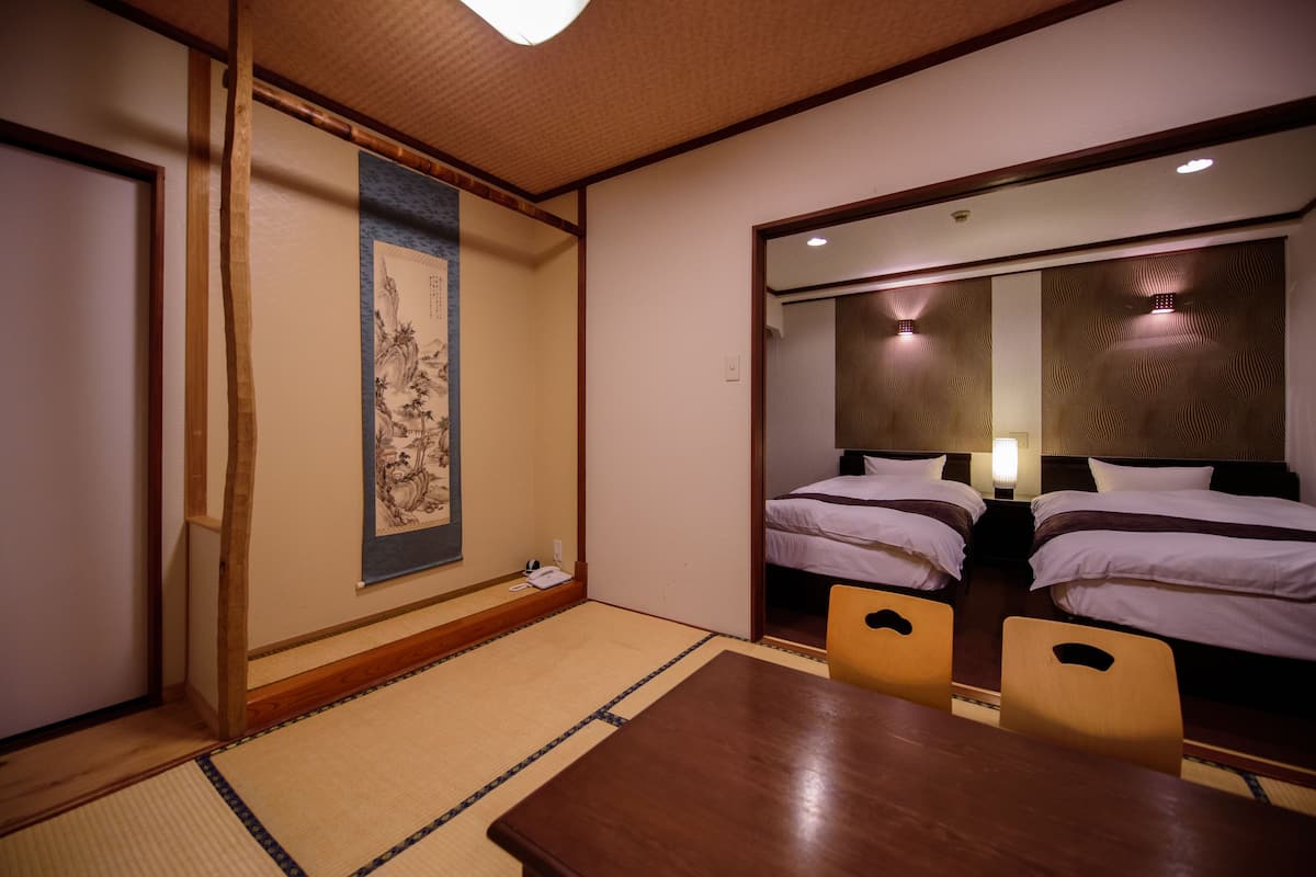 A traditional Japanese hotel room with tatami flooring, a wooden table, two beds, and a large mirror reflecting the room.