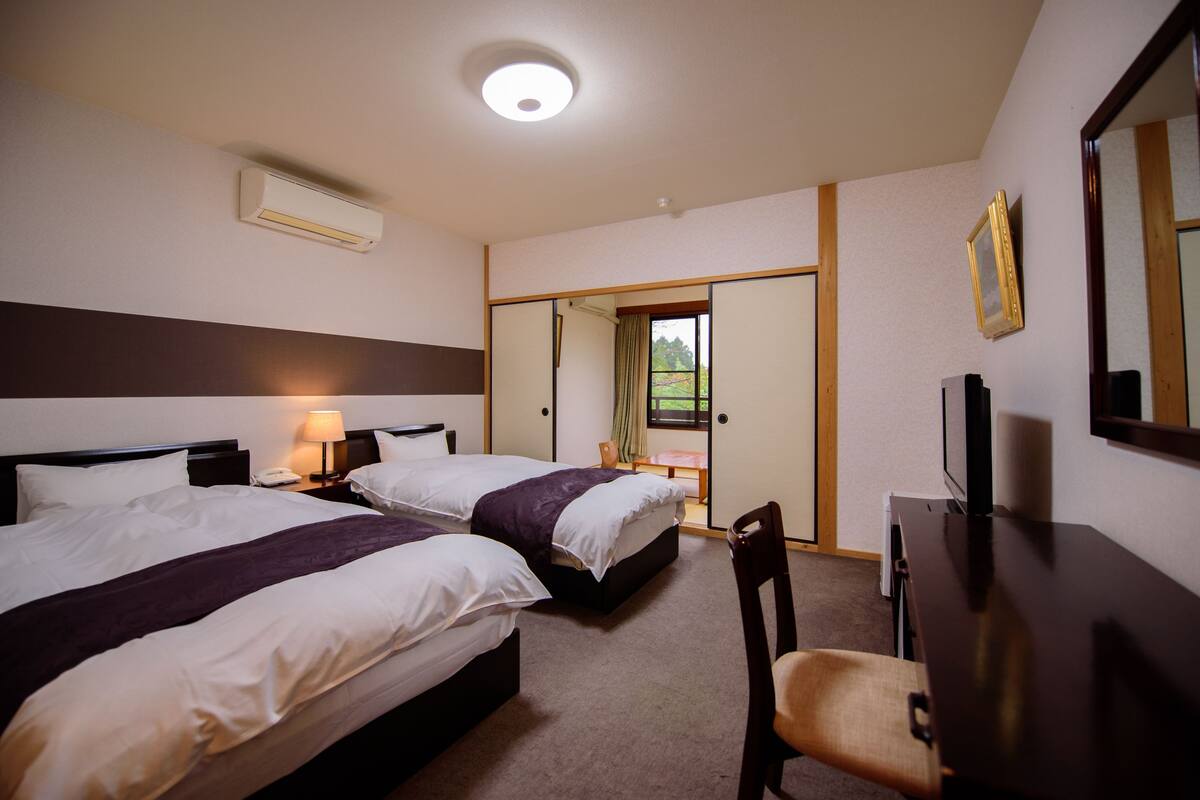 A hotel room with two beds, a desk, a chair, a TV, and a sliding door leading to a balcony.