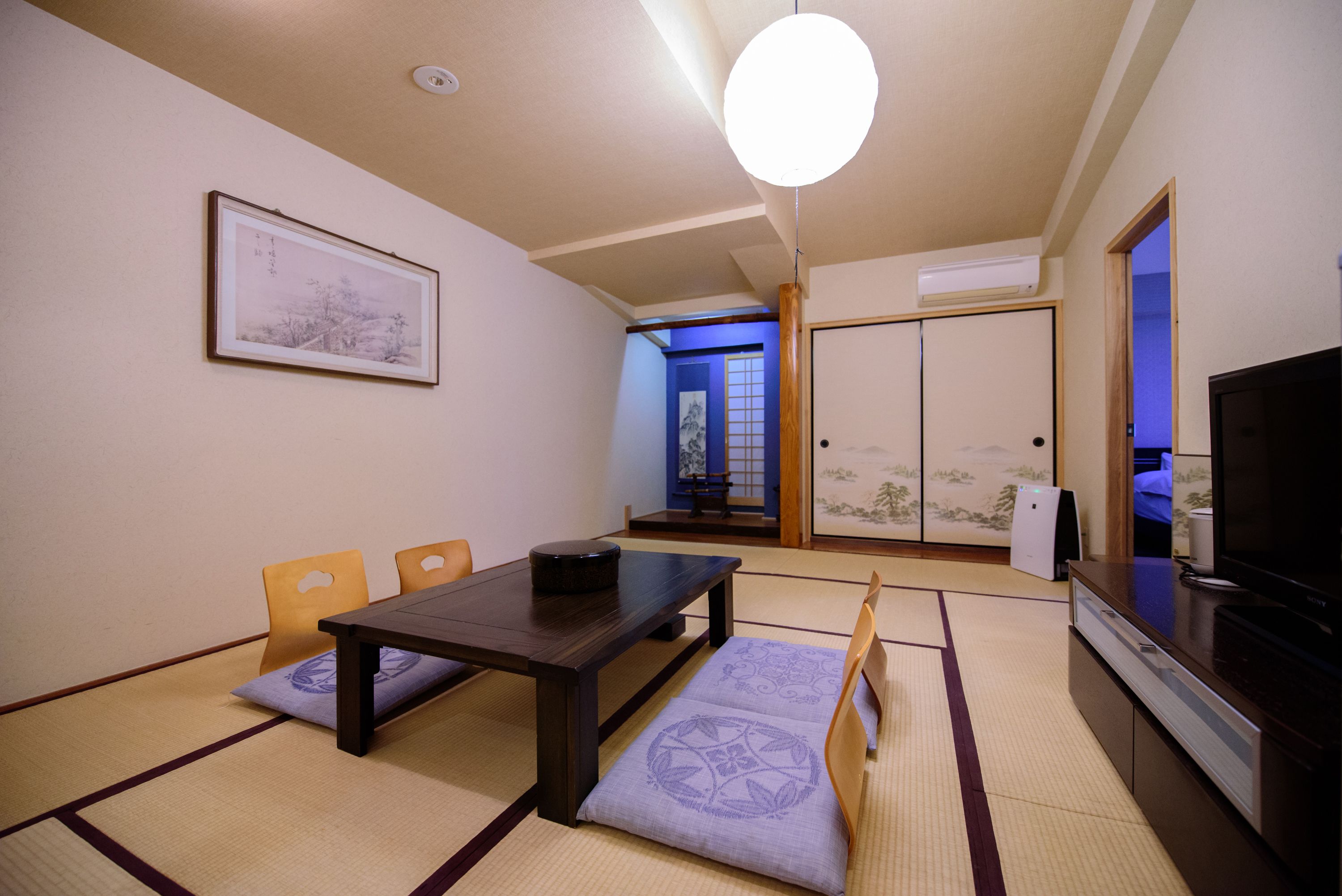 A traditional Japanese-style room with tatami flooring, a low table, cushions, a television, and a sliding door leading to another room.