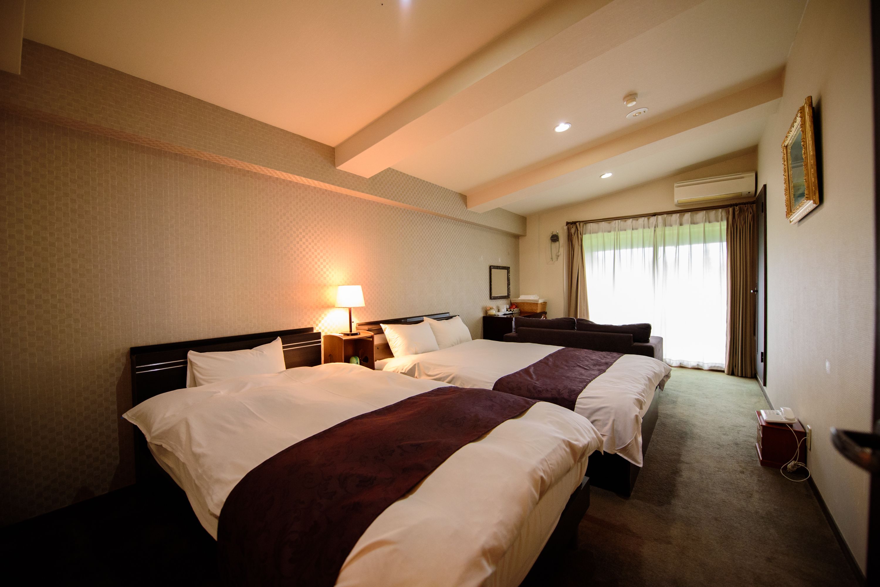 Quarto de hotel com duas camas, uma mesa de cabeceira com luminária, uma escrivaninha com cadeira e uma janela com cortinas.