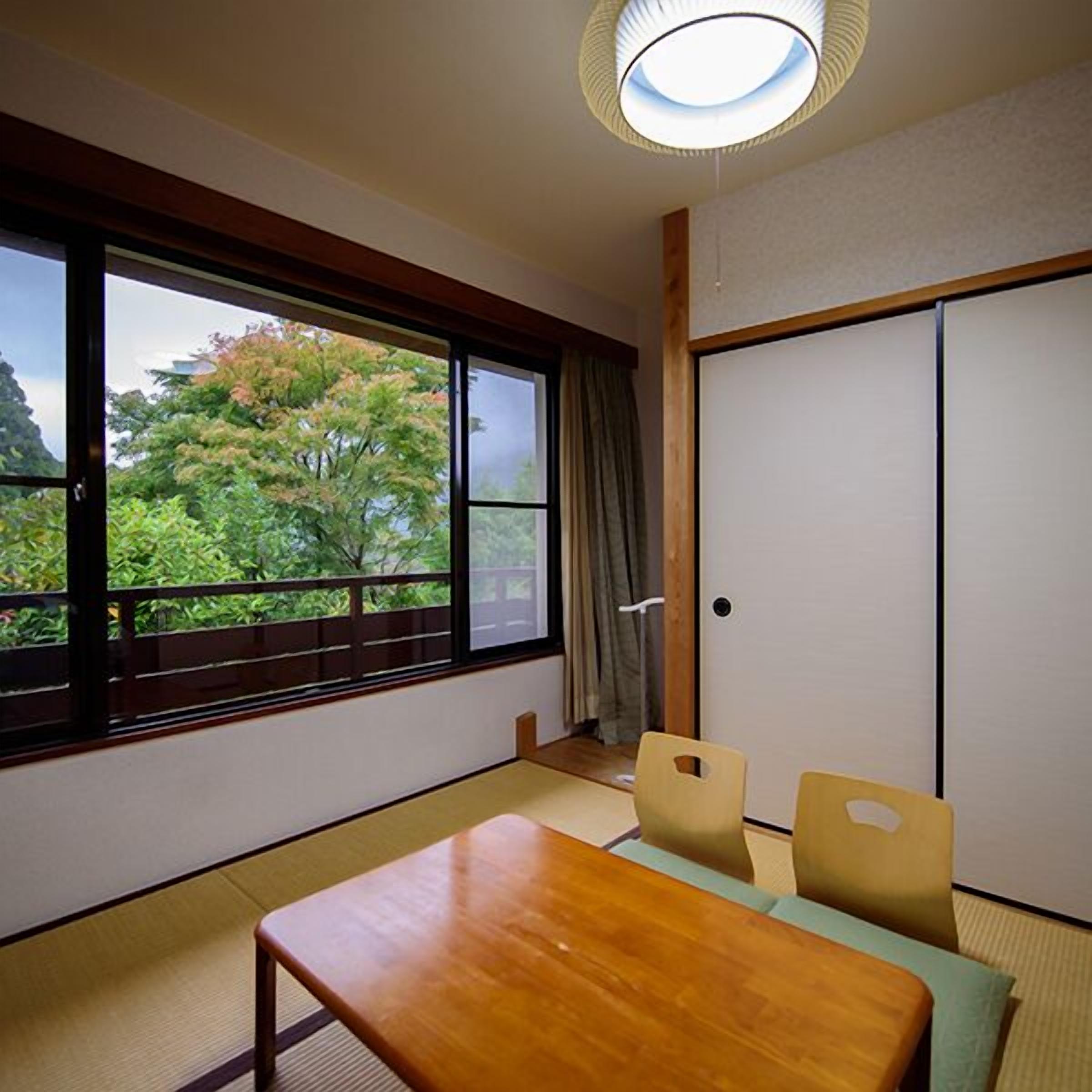 A room with tatami flooring, a wooden table, two chairs, a sliding door, and a view of trees through a large window.