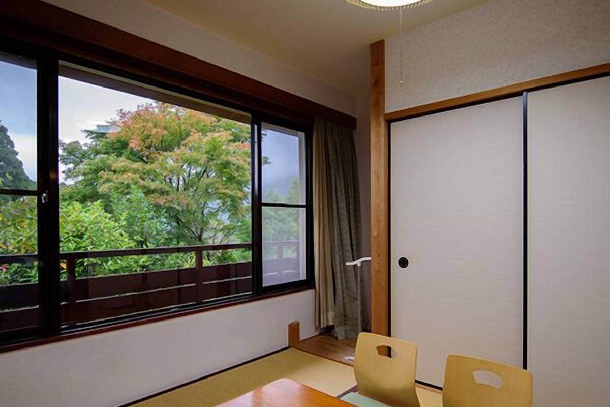 A room with tatami flooring, a wooden table, two chairs, a sliding door, and a view of trees through a large window.