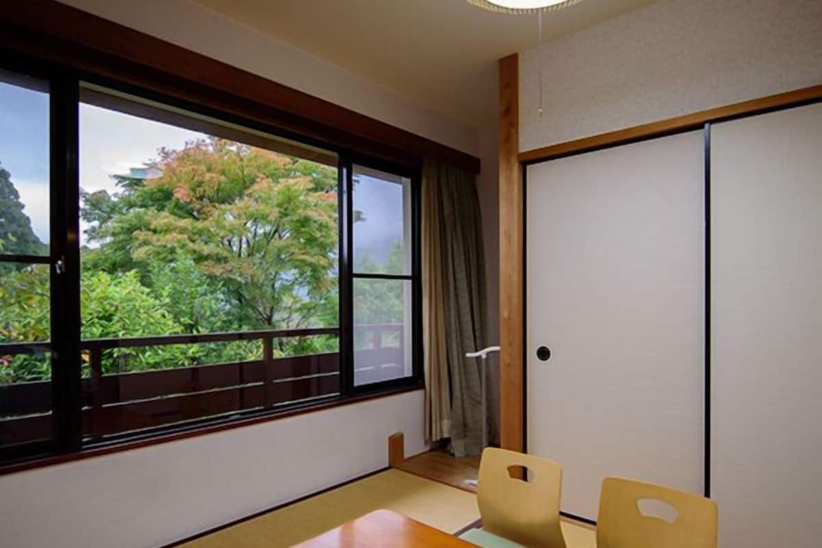 Una habitación con suelo de tatami, una mesa de madera, dos sillas, una puerta corredera y vistas a árboles a través de un ventanal grande.