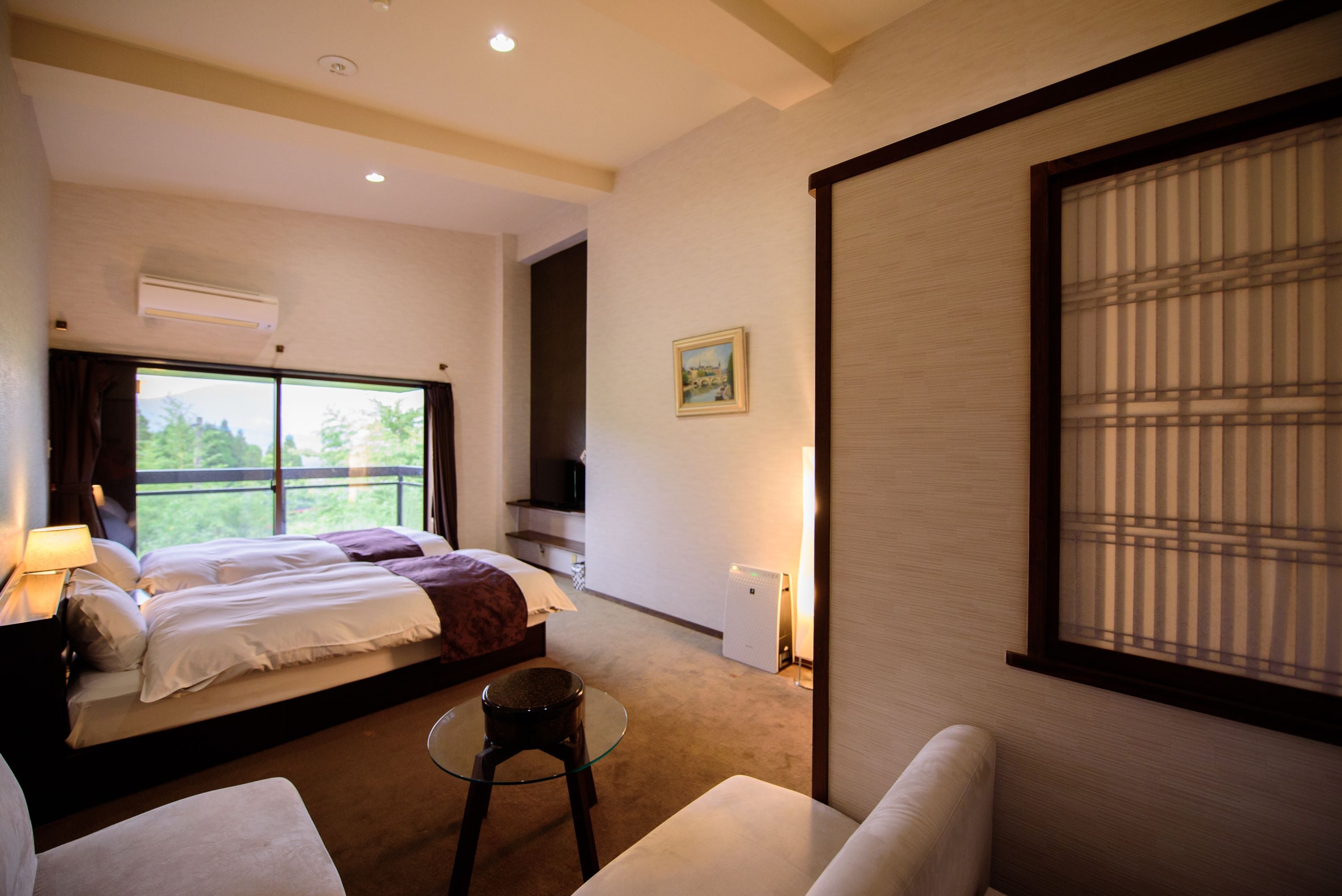 A hotel room with a bed, a small table, a sofa, a TV, and a window with blinds.