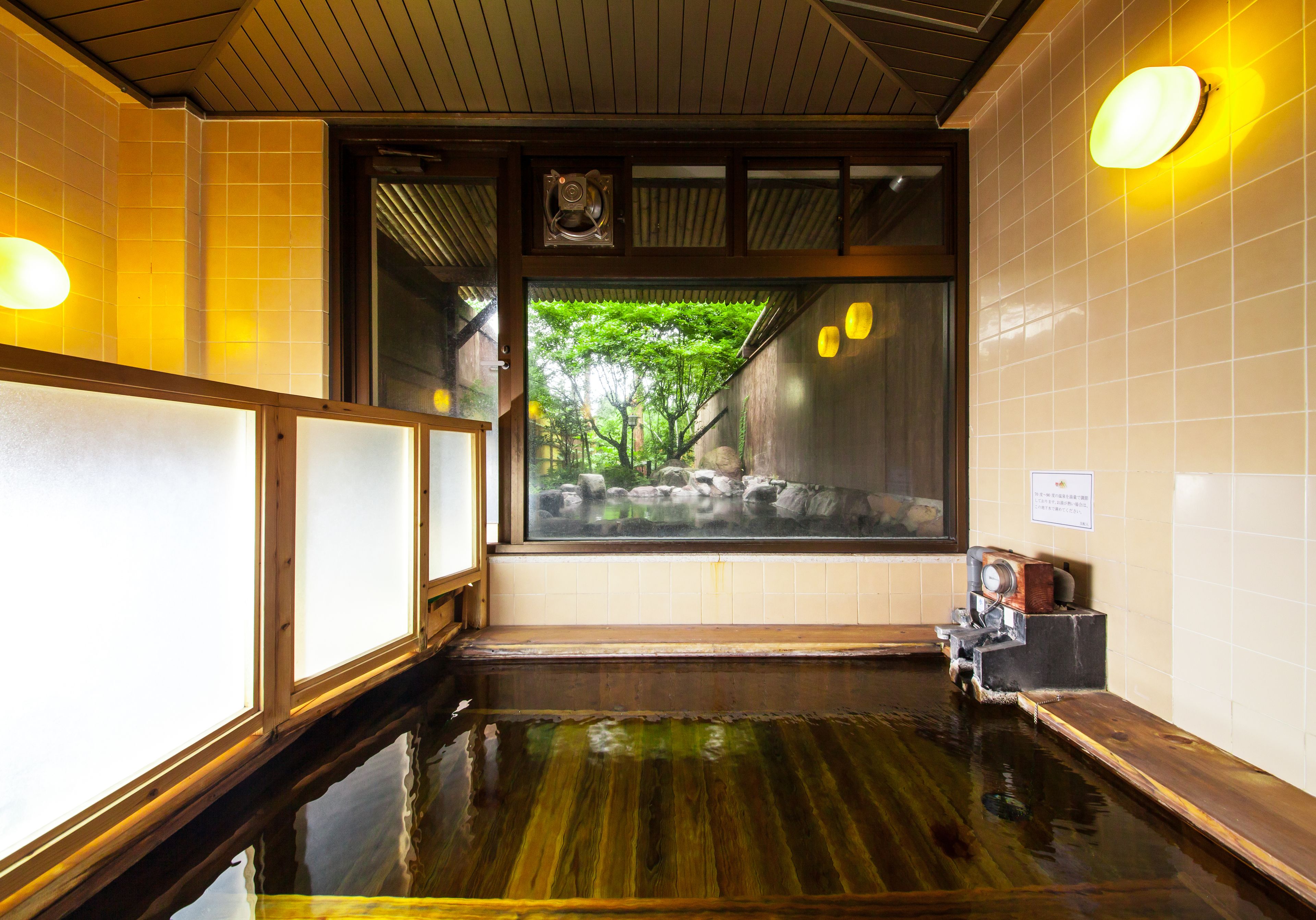 A traditional Japanese onsen with a wooden interior, a view of trees through a glass window, and a stone bath area.