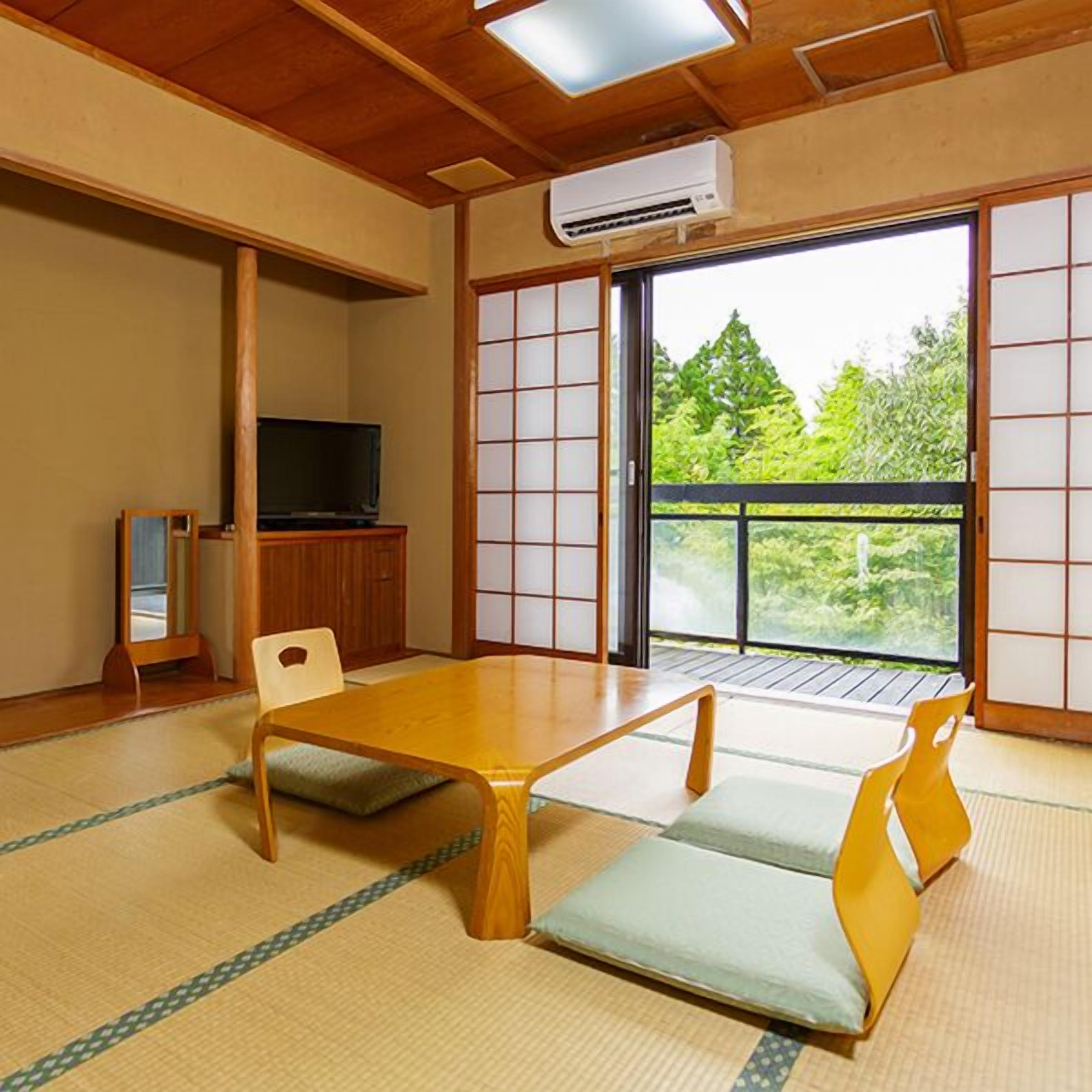 Um quarto japonês tradicional com tatames, uma mesa baixa, uma televisão e portas deslizantes que dão para uma varanda com vista para o verde.