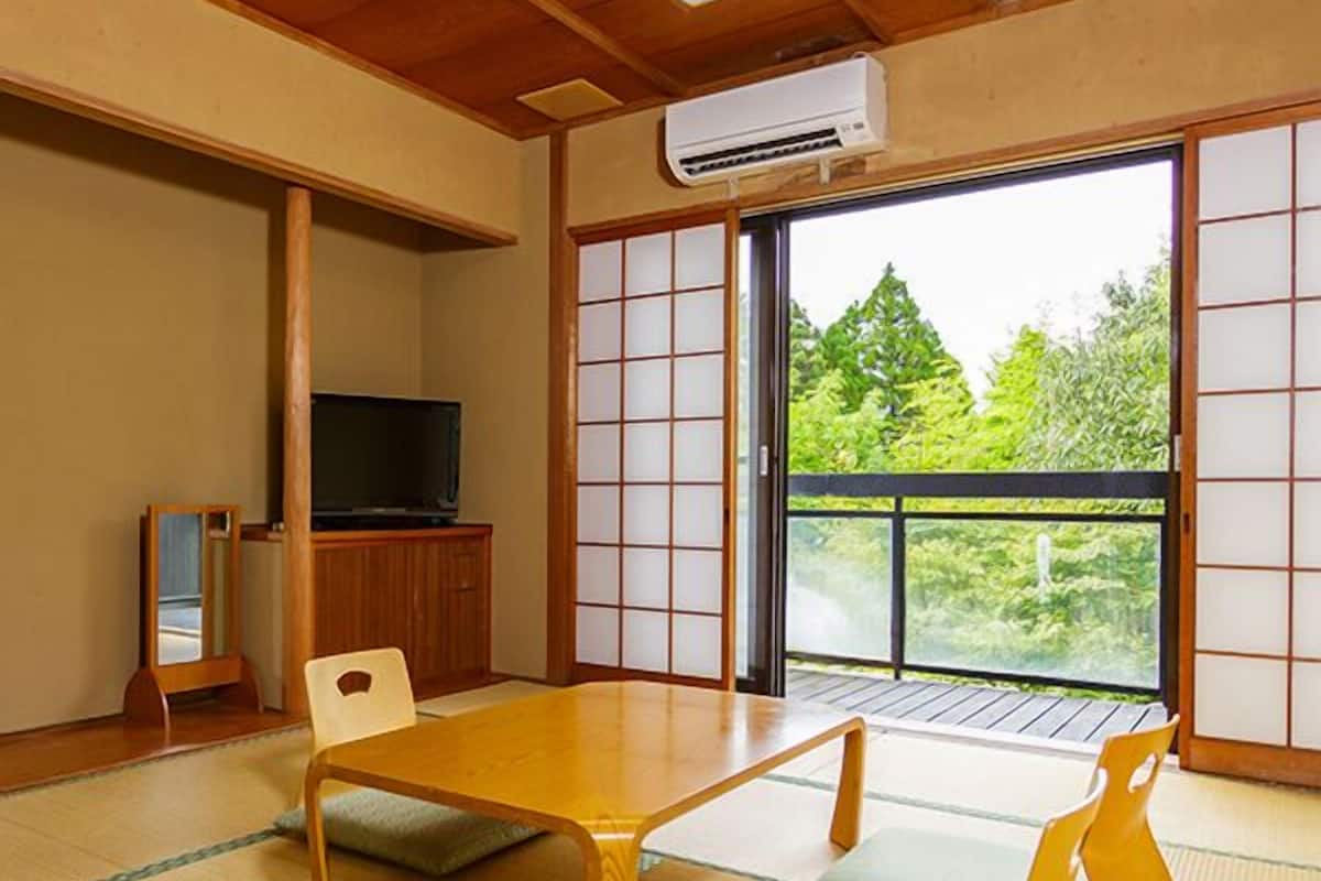 Ett traditionellt japanskt rum med tatamimattor på golvet, ett lågt bord, en tv och skjutdörrar som leder ut till en balkong med utsikt över grönskan.