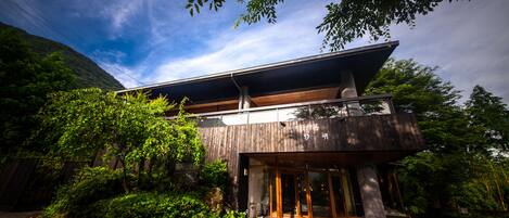 A modern building with a sloped roof and large glass doors, surrounded by lush greenery and a mountainous backdrop.