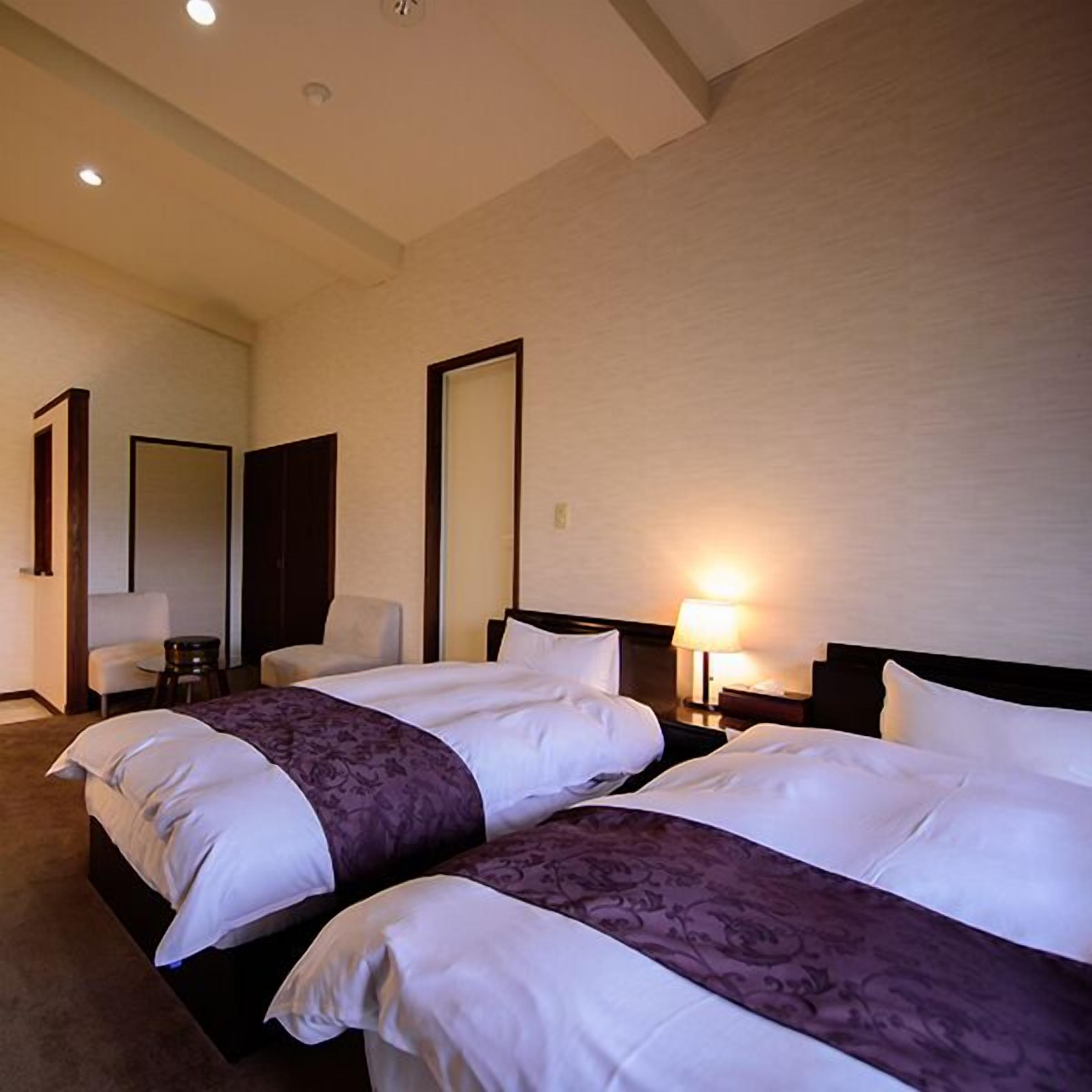 Quarto de hotel com duas camas, um espaço de estar com uma poltrona e uma mesa pequena, e um espelho na parede.
