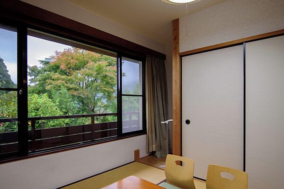 A room with tatami flooring, a wooden table, two chairs, a sliding door, and a view of trees through a large window.