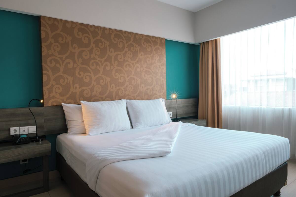 A neatly made bed in a hotel room with a patterned headboard, a bedside table with a lamp, and a window with curtains.