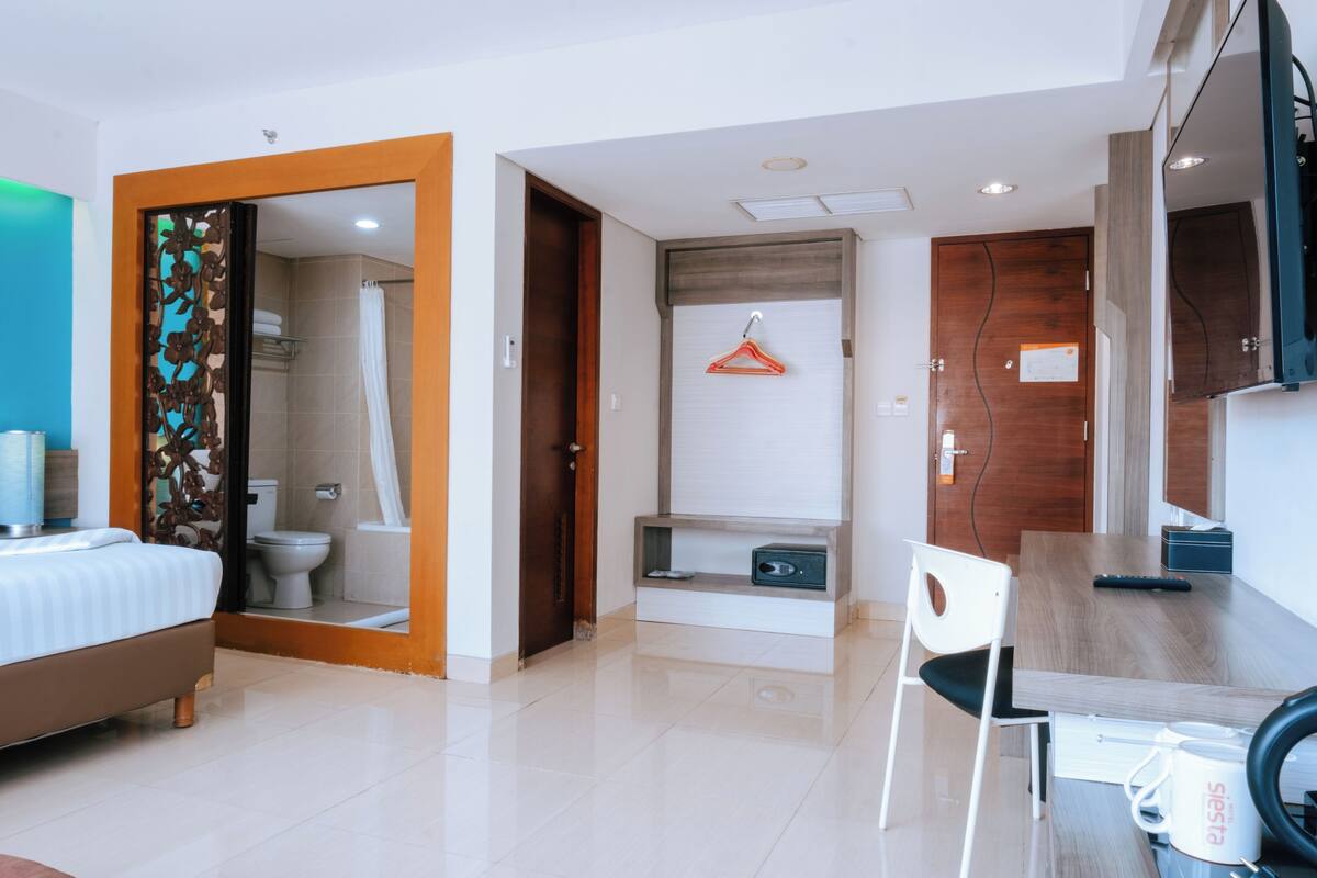 A hotel room with a bed, a desk, a chair, a hanging clothes rack, and a shower area.