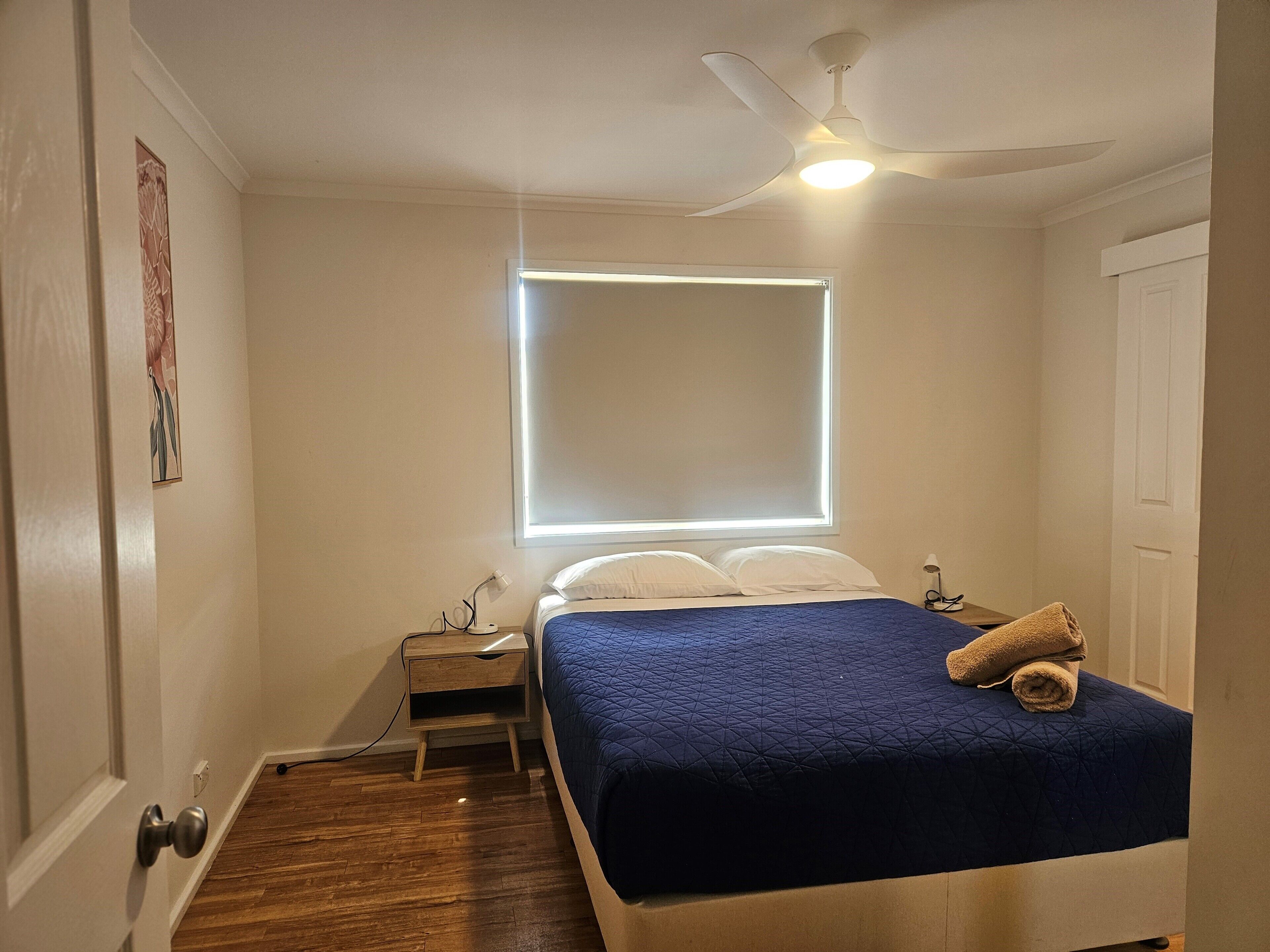 Une chambre à coucher comprenant un lit, une table de chevet, un ventilateur de plafond et une fenêtre avec des stores.