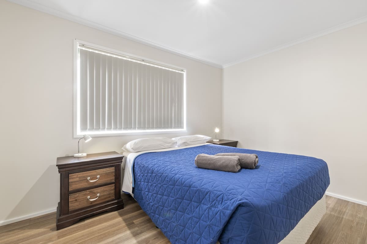 Une chambre avec un grand lit, une table de chevet avec des tiroirs, une lampe de chevet et une fenêtre avec des stores.