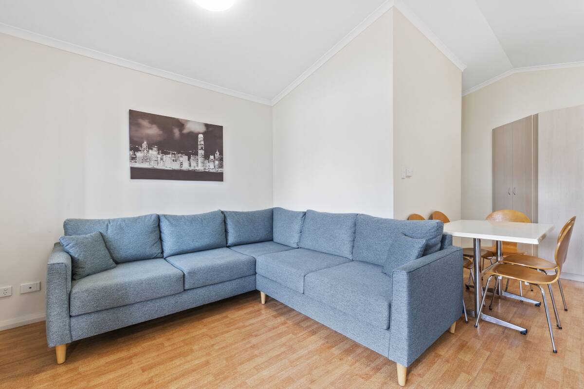 Un salon moderne avec un canapé d’angle gris, une table à manger blanche avec des chaises, et une œuvre d’art représentant un paysage urbain accrochée au mur.