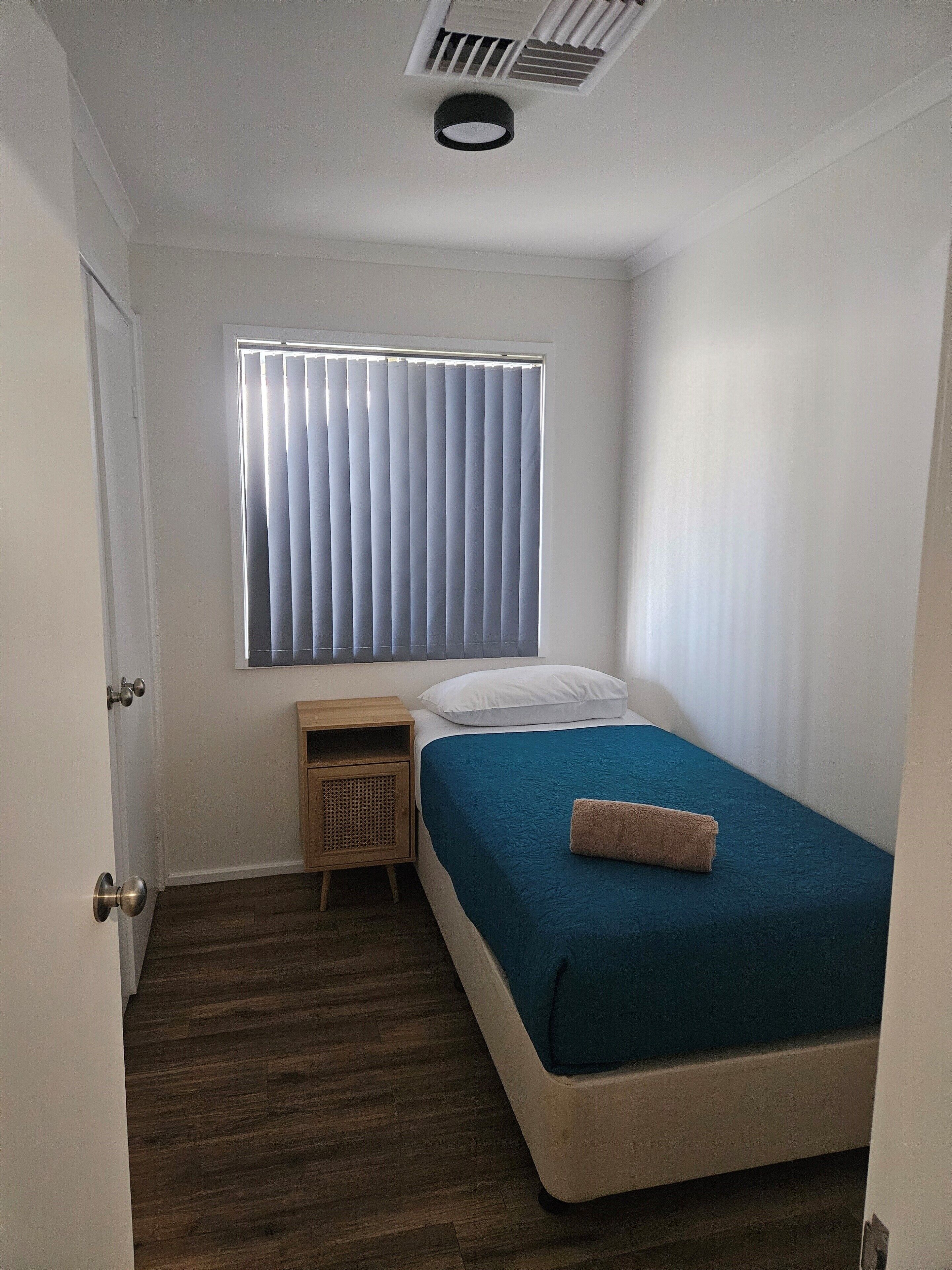 Un dormitorio pequeño y ordenado con una cama individual, una mesita de noche, una ventana con persianas y un respiradero en el techo.