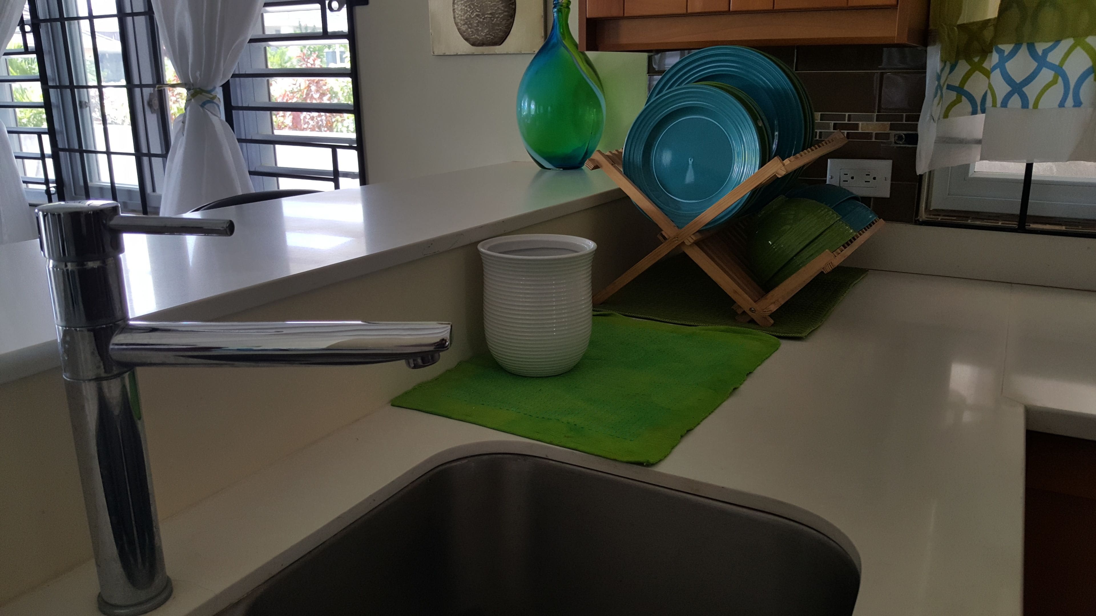 Una cocina con un fregadero de acero inoxidable, un tapete verde y un escurreplatos con platos azules.