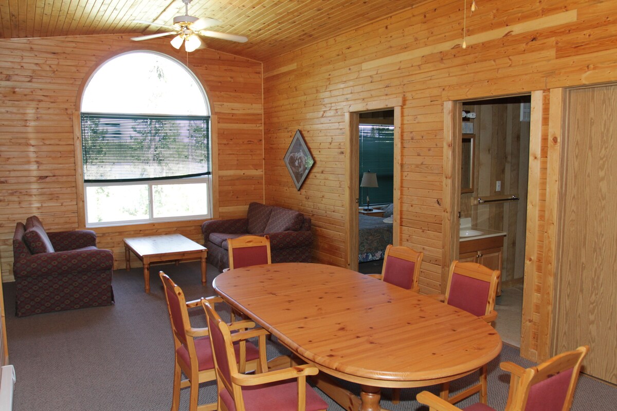 A wooden cabin interior with a dining table, chairs, a sofa, and a window with blinds.