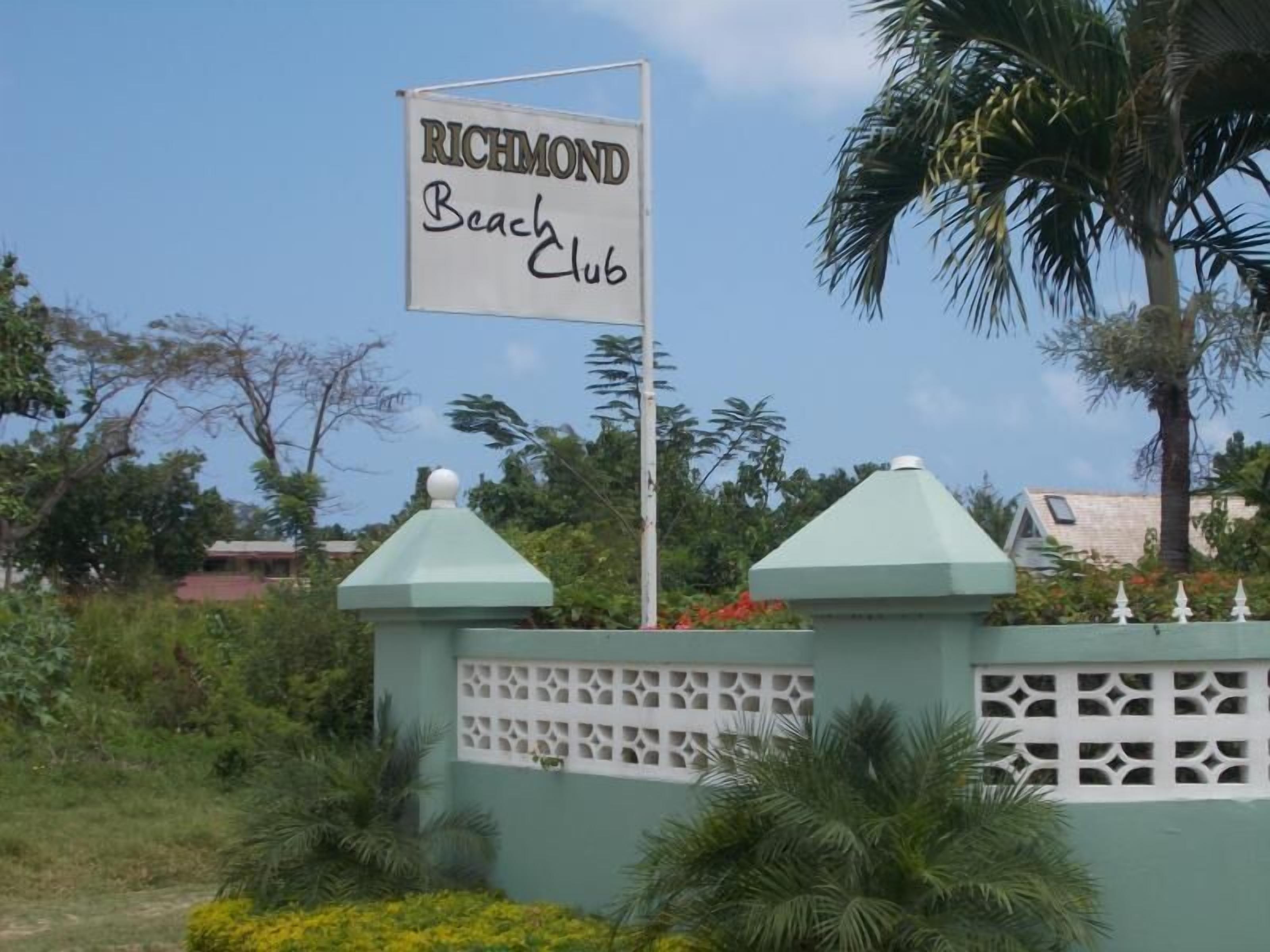 A sign for Richmond Beach Club with a green fence and palm trees in the background.