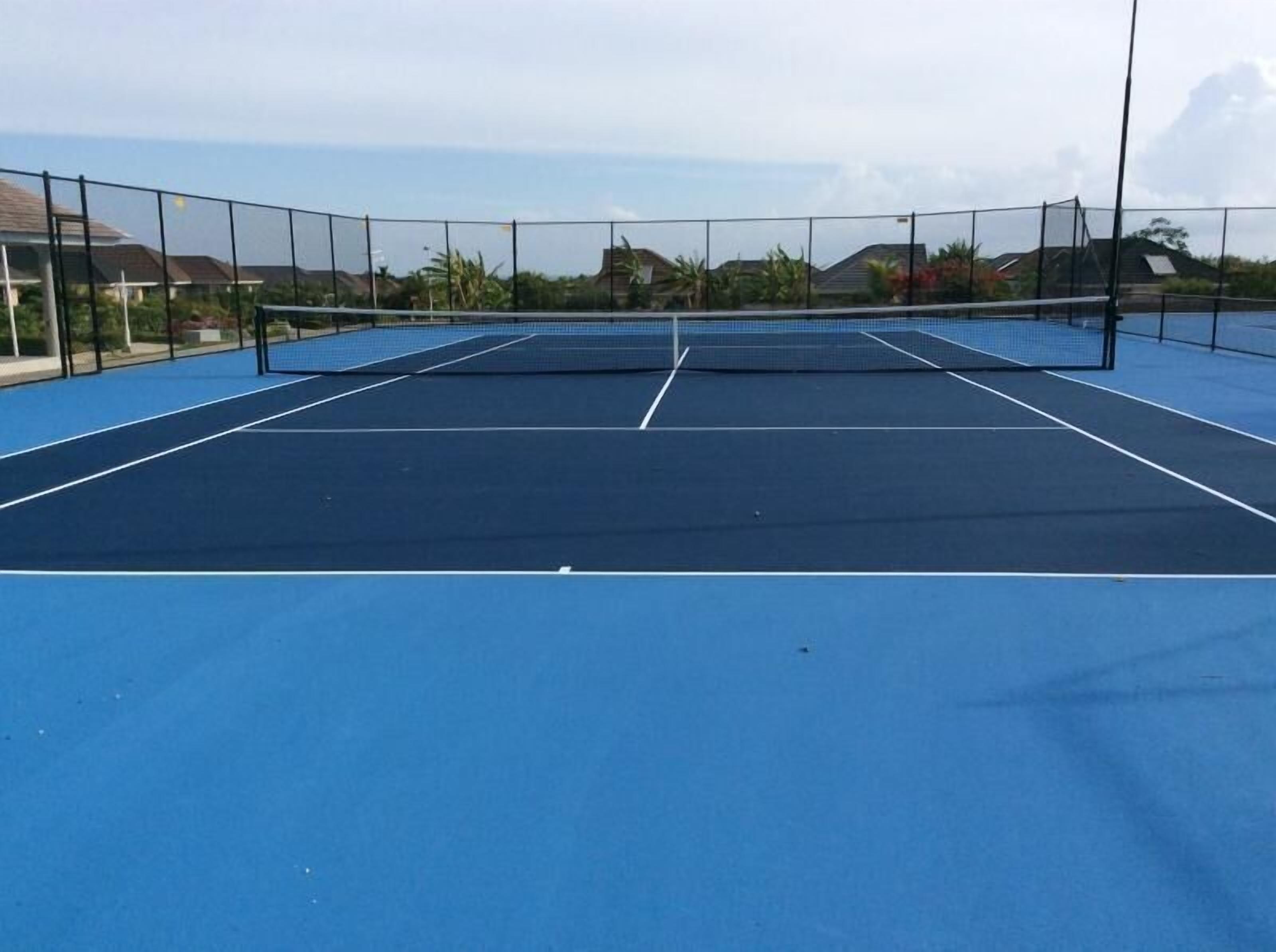 Un court de tennis en plein air, doté d’un filet et entouré d’une clôture, avec des maisons en arrière-plan.